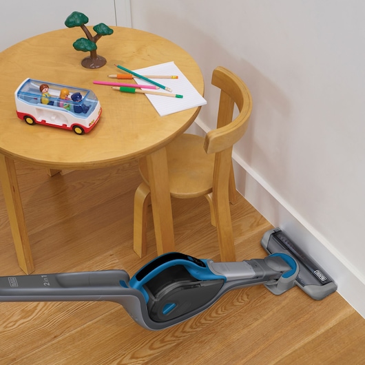 A BLACK+DECKER 2-in-1 cordless stick vacuum cleaner is cleaning along the edge of a wooden floor near a small round wooden table and chair. The table has colored pencils, a notepad, a toy bus with figures, and a small toy tree on it.