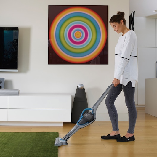 A person with a blurred face uses a BLACK+DECKER cordless stick vacuum cleaner (model SVJ520BFS) to clean a green rug in a modern living room. The room features a colorful circular wall art, a white cabinet, and wooden flooring.