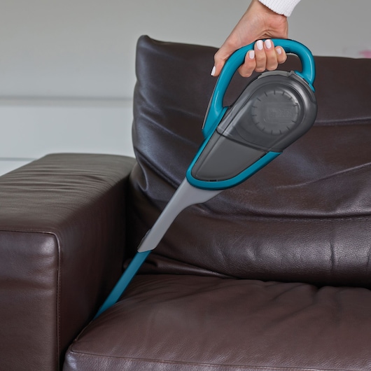 A close-up of a hand holding a BLACK+DECKER handheld vacuum cleaner, cleaning between cushions of a brown leather sofa.