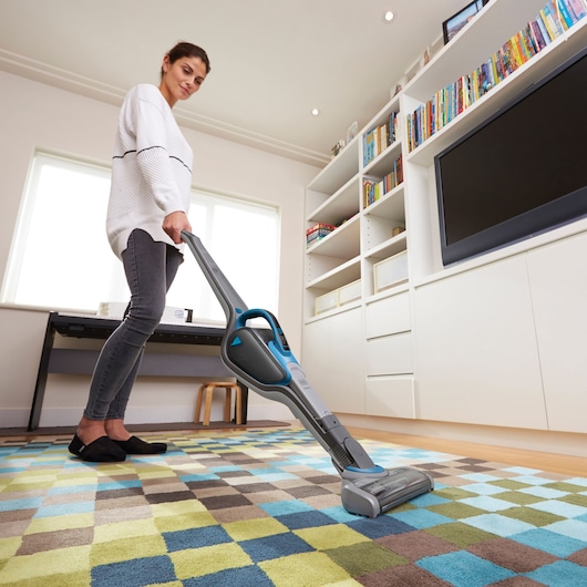A person vacuuming a colorful checkered rug in a modern living room with a BLACK+DECKER SVJ520BFS cordless vacuum. The room has white shelving, a large television, and natural light coming in from the windows.