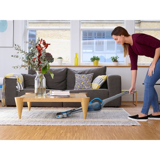 A person uses a BLACK+DECKER stick vacuum cleaner (model SVJ520BFS) to clean under a wooden coffee table in a modern living room with a gray sofa, decorative pillows, and plants on the windowsill.