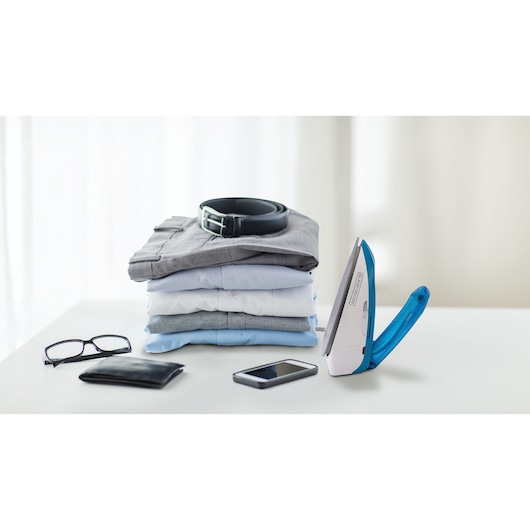 A compact BLACK+DECKER iron with a blue handle stands on a white surface next to a neatly folded stack of shirts, a black belt on top, glasses, a wallet, and a smartphone. Bright, soft background with curtains.