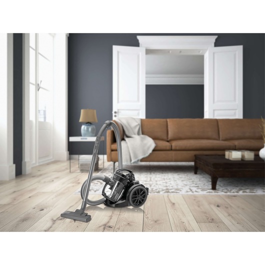 A BLACK+DECKER canister vacuum cleaner is placed on a light wood floor in a modern living room with a brown sofa, dark accent wall, white doors, and contemporary décor.