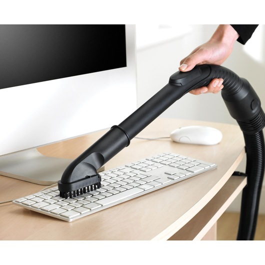 A person uses a vacuum cleaner with a brush attachment to clean a computer keyboard on a desk. A computer monitor and mouse are also visible.