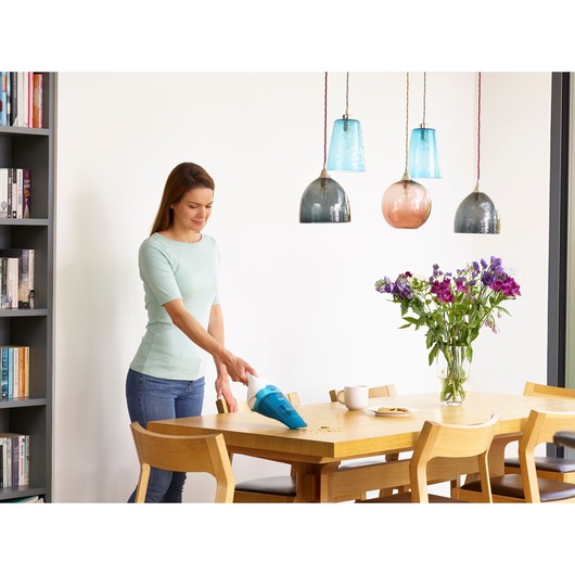 A person using a handheld BLACK+DECKER vacuum cleaner (model WDC115WA) to clean crumbs from a wooden dining table in a bright, modern room. There are flowers in a vase and a cup on the table.