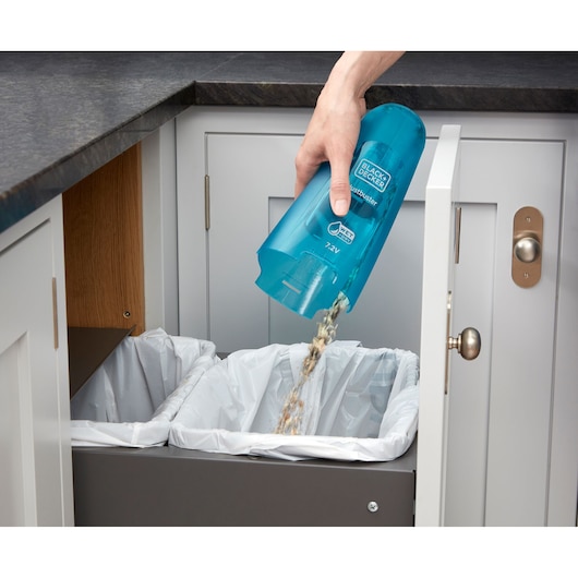 A person is emptying the contents of a BLACK+DECKER handheld vacuum cleaner into a kitchen trash bin lined with white plastic bags.